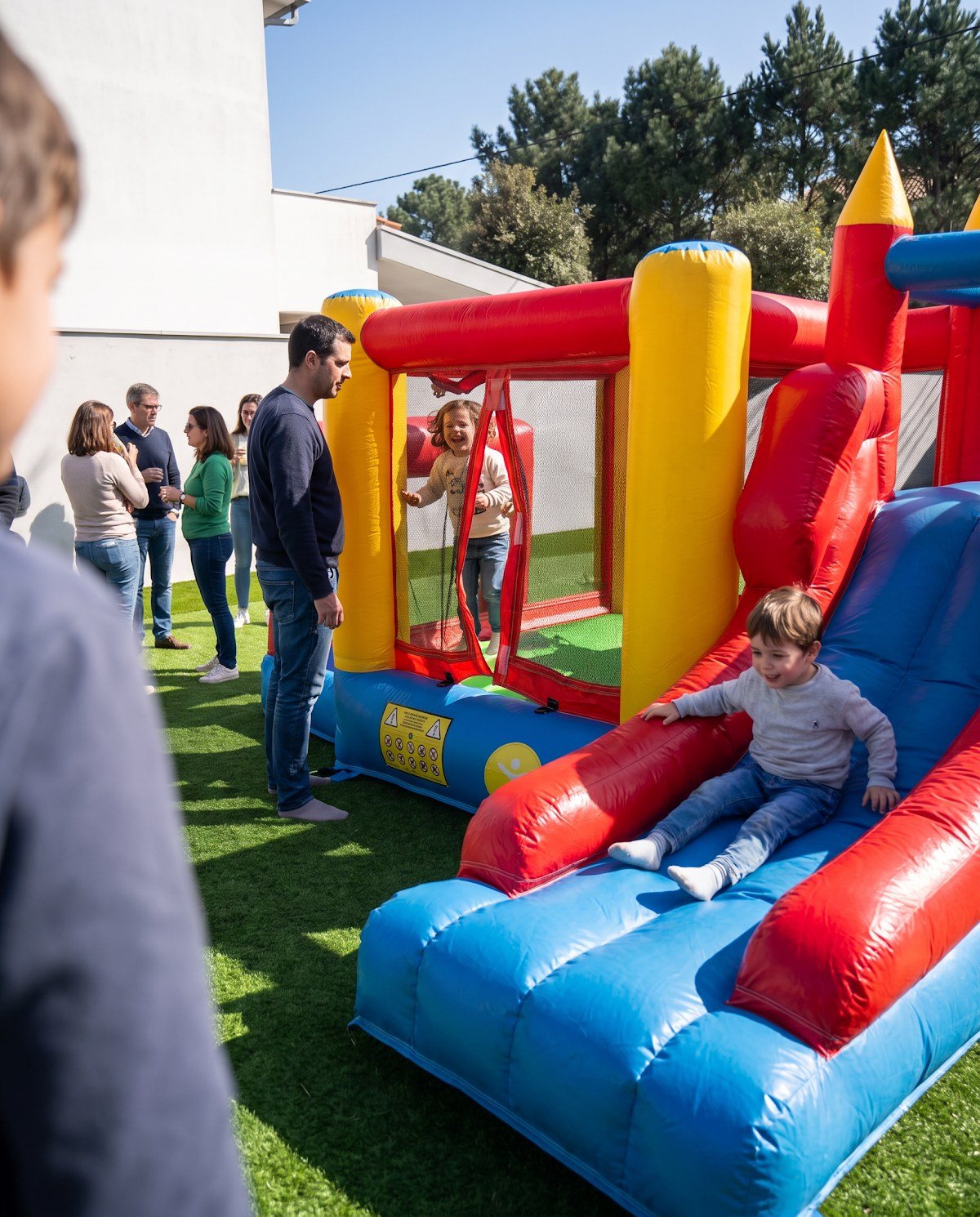 Crianças a brincar num insuflável colorido num jardim, com convidados ao lado